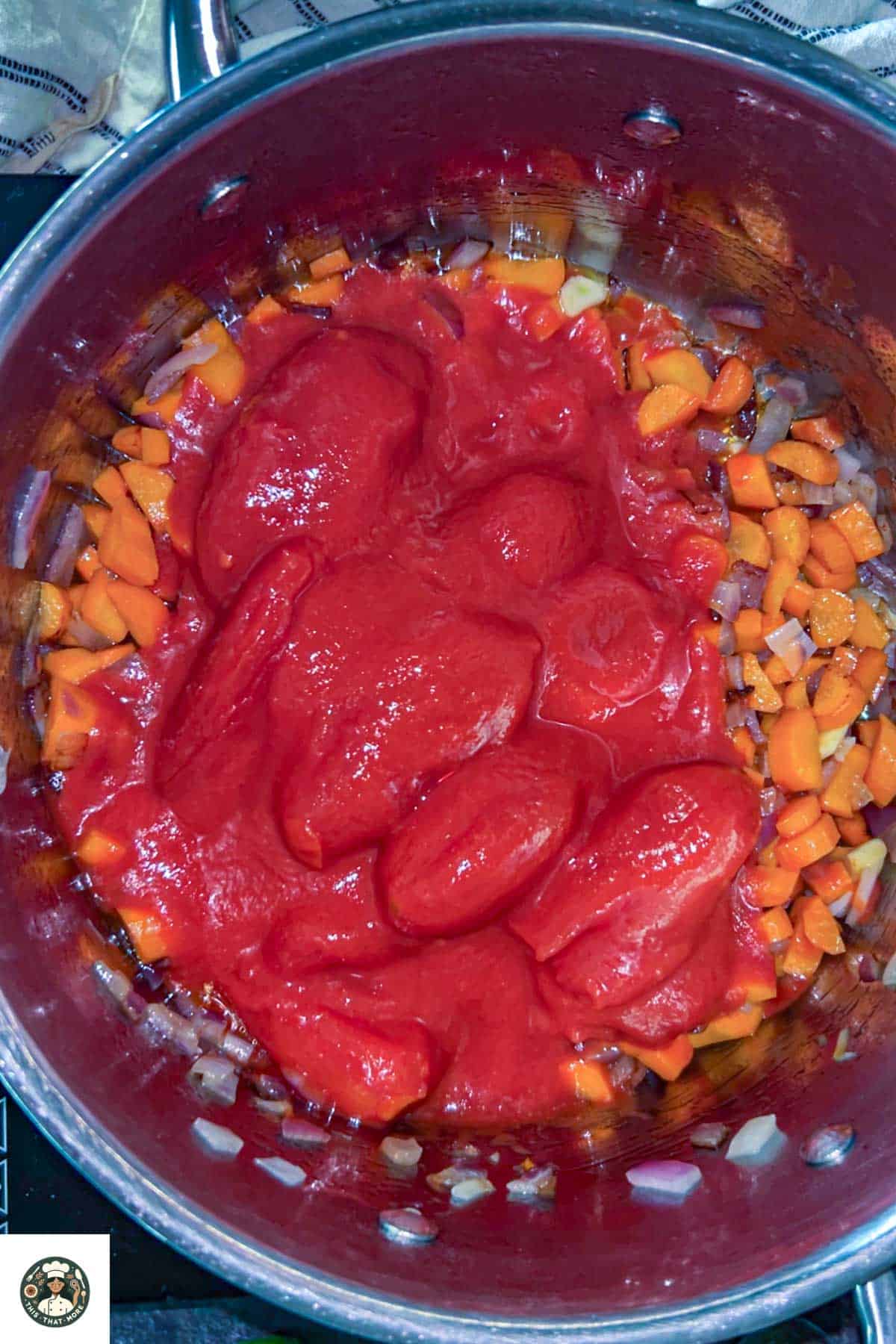 Image of large steel pot with caramelized onion, garlic, and carrot topped with canned tomatoes.