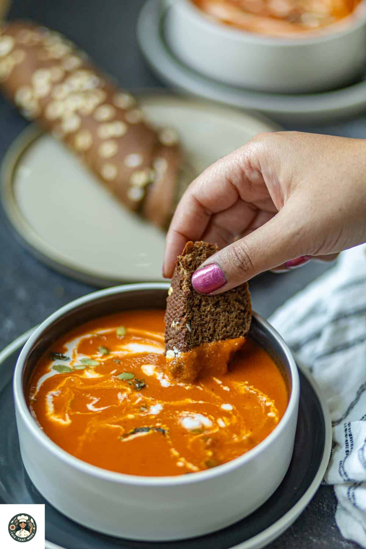 A brown bread dipped in dair-free tomato soup served in a white bowl with garnish of coconut milk, basil, and pumpkin seeds.