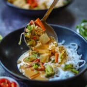 Close-up of creamy Thai curry with tofu and vegetables being served over rice noodles.