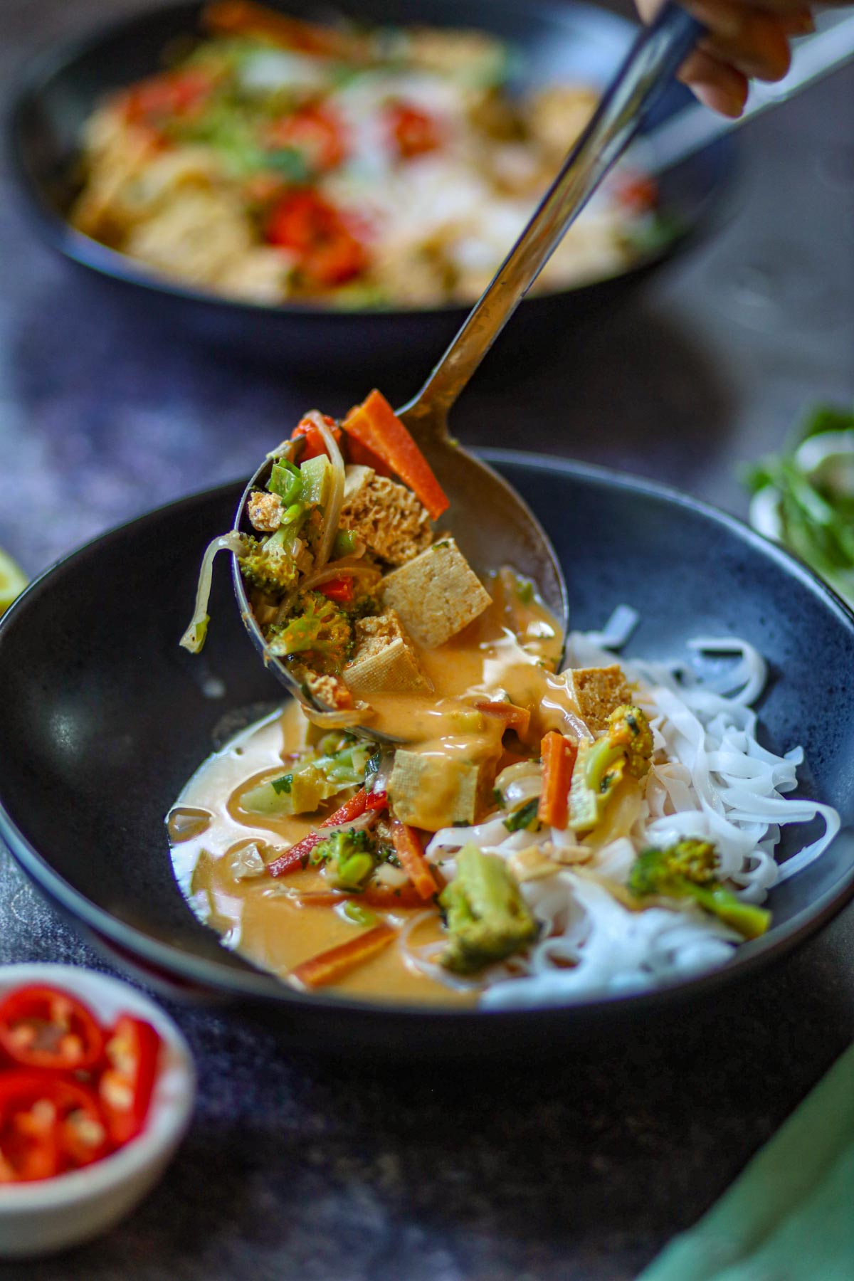 Close-up of creamy Thai curry with tofu and vegetables being served over rice noodles.
