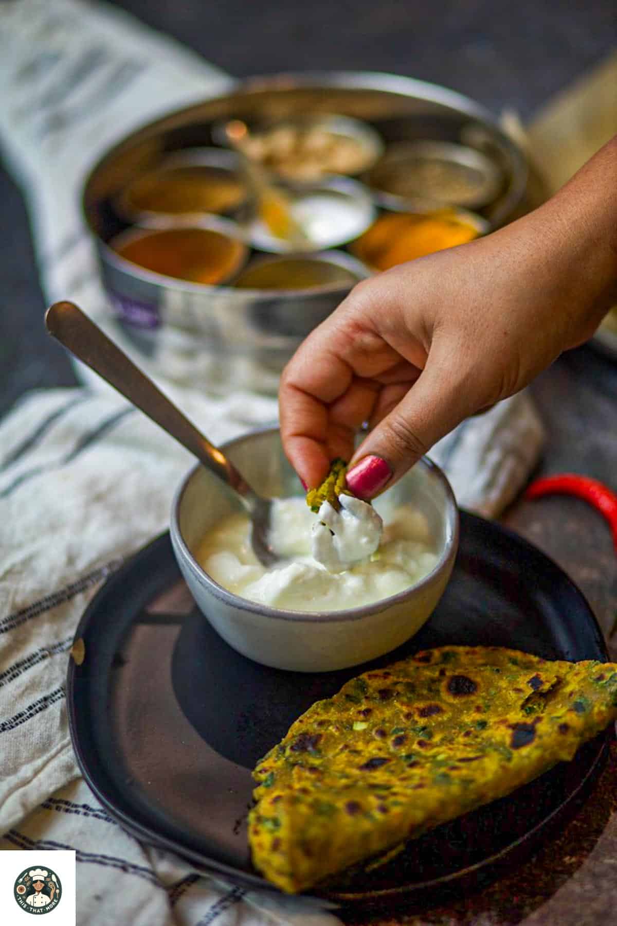 Image of a bite of methi paratha dipped in yogurt with the whole paratha on the black plate.