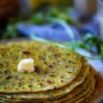 Side shot of stack of methi paratha rcipe on a white plate with cilantro showing at the background.
