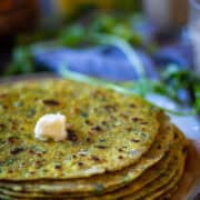 Side shot of stack of methi paratha rcipe on a white plate with cilantro showing at the background.