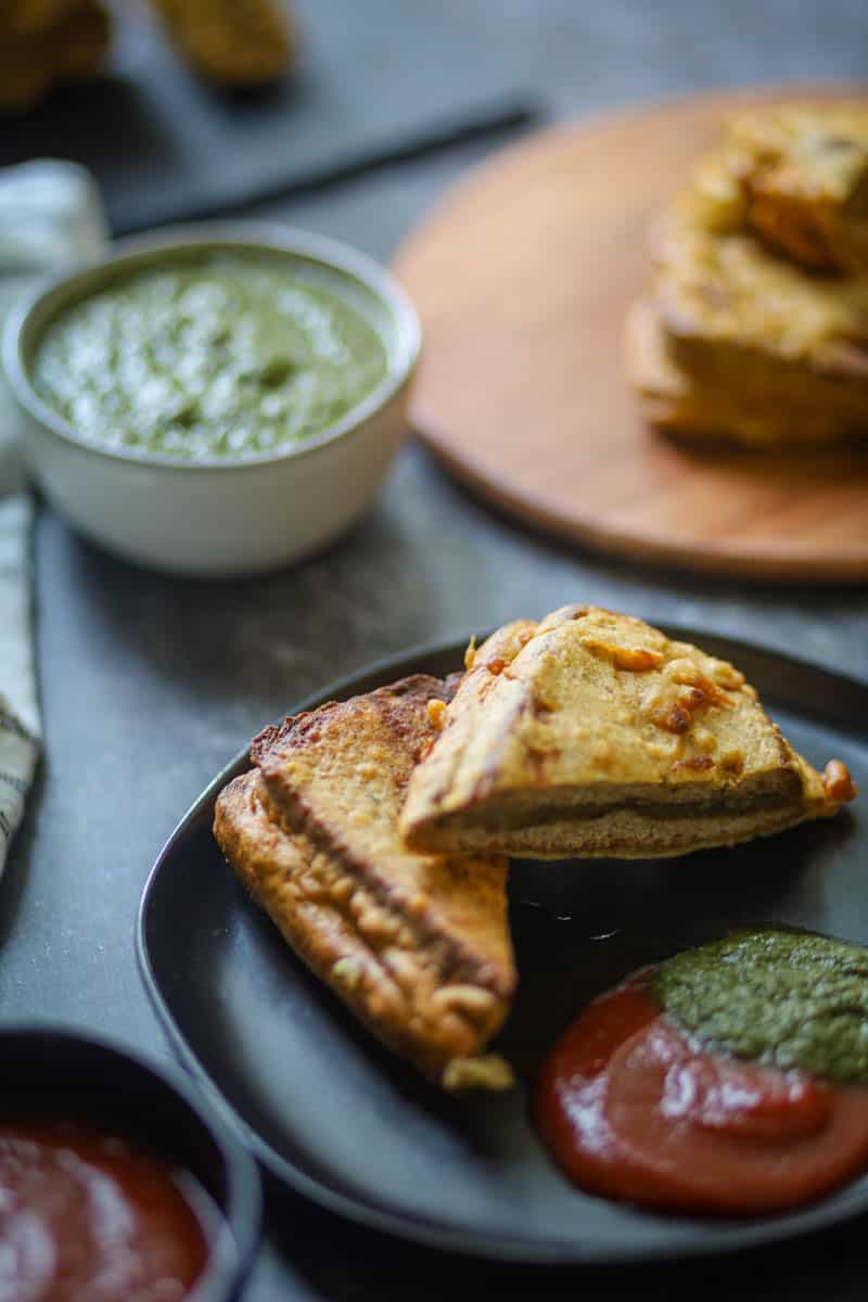 Bread Pakora served on a black plate with green chutney and ketchup.