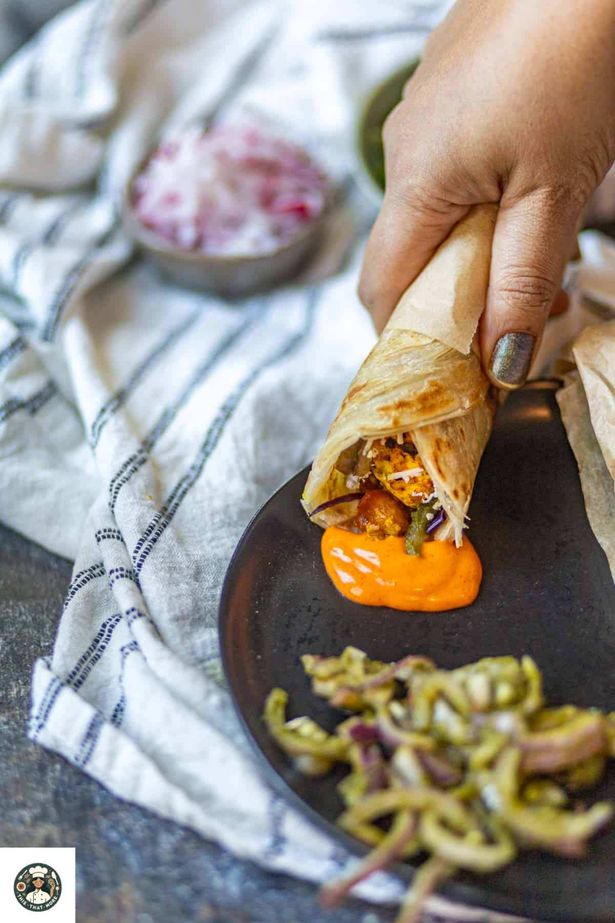 Paneer kathi roll dipped in hot sauce on a black plate.