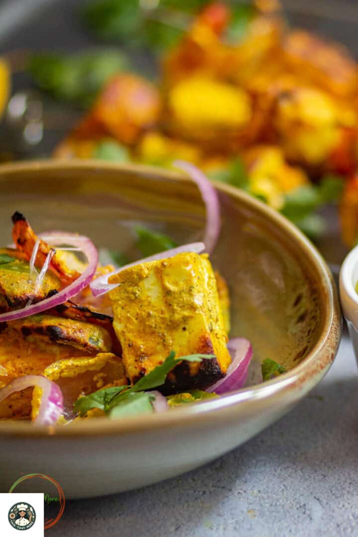 Side shot of tandoori paneer tikka in an olive green bowl served with sliced onions and baking tray kept on the side over a white background. 