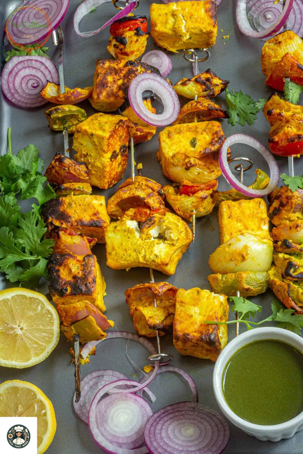 Image showing grilled and charred tandoori paneer tikka in a baking tray served with green chutney in sliced red onions.