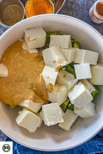 Image showing yogurt marination is added to onion, bell peppers, and paneer in a white bowl.,