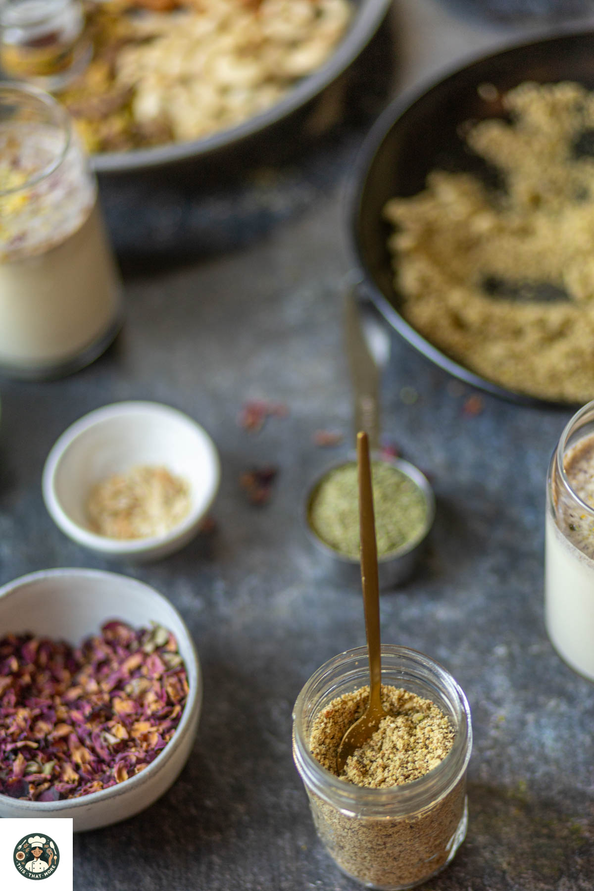 Homemade thandai powder in a mason jar surrounded by almonds, fennel seeds, and rose petals on a tray.