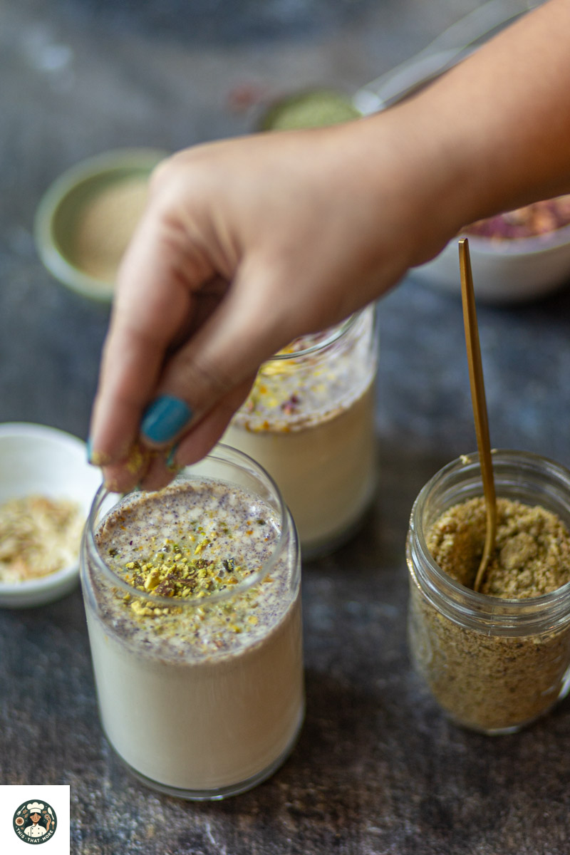 Hand sprinkling homemade thandai powder over glass filled with thandai milk.