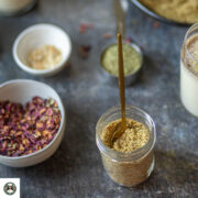 Homemade thandai powder in a mason jar surrounded by almonds, fennel seeds, and rose petals on a tray.