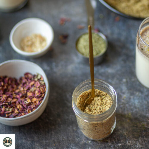 Homemade thandai powder in a mason jar surrounded by almonds, fennel seeds, and rose petals on a tray.