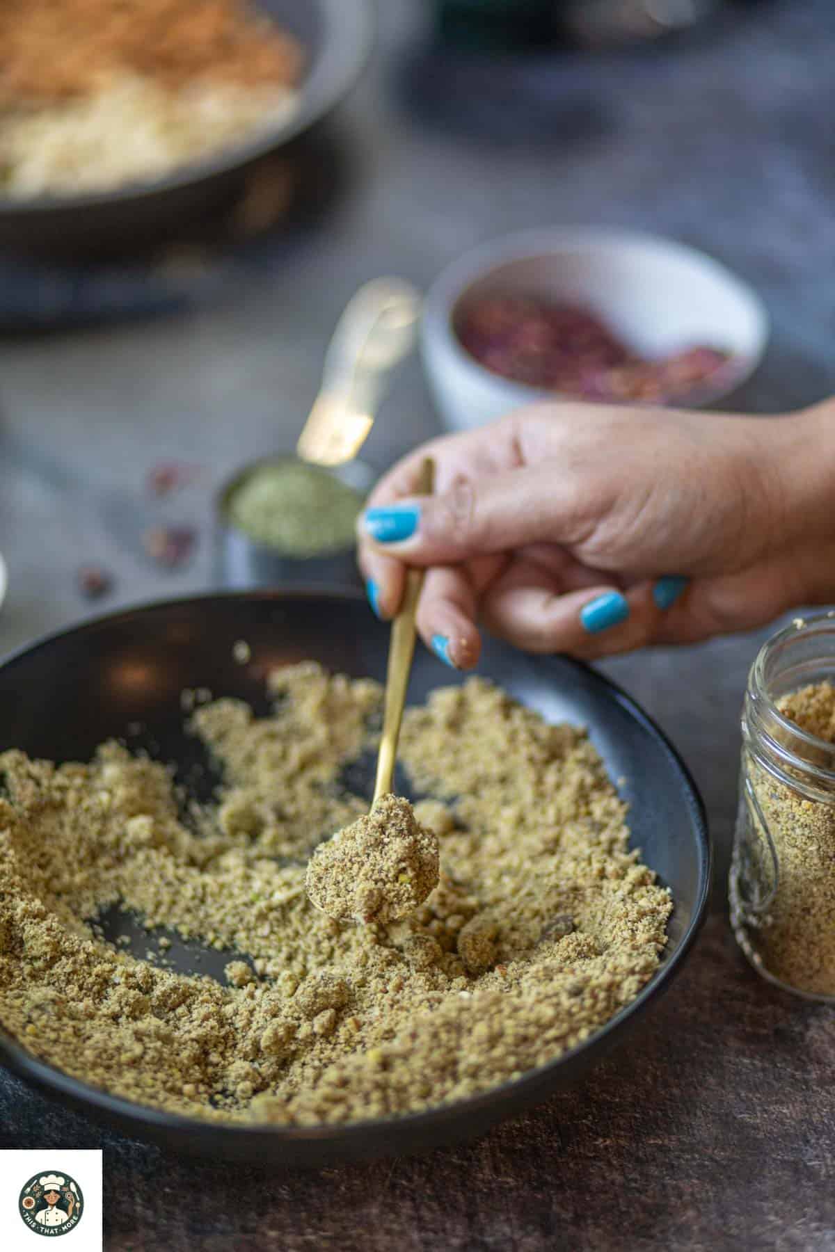 Image showing a spoonful of thandai powder in a black bowl.