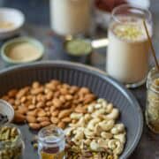 Image showing nuts an daromatic spices placed in a gray tray with thandai served in tall glasses and placed at the back of the gray tray.