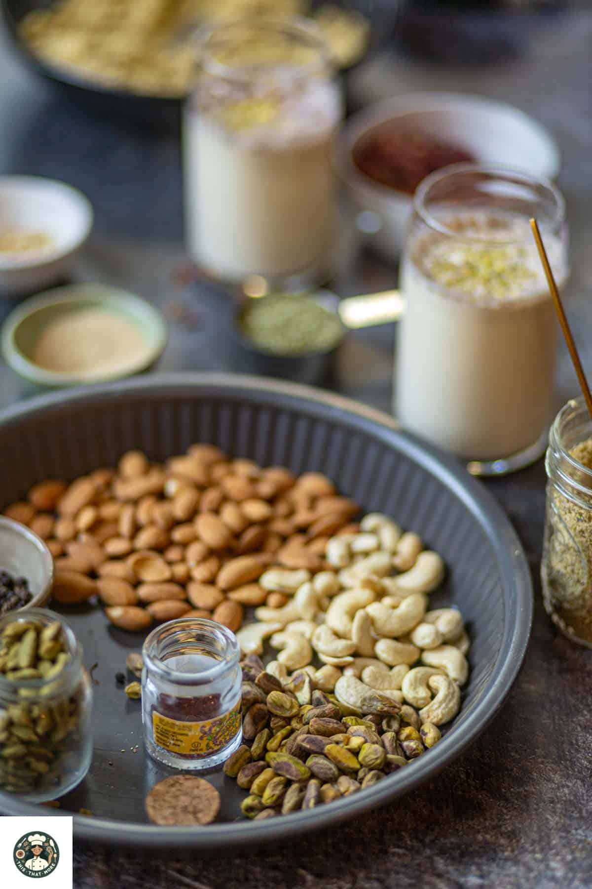 Image showing nuts an daromatic spices placed in a gray tray with thandai served in tall glasses and placed at the back of the gray tray.