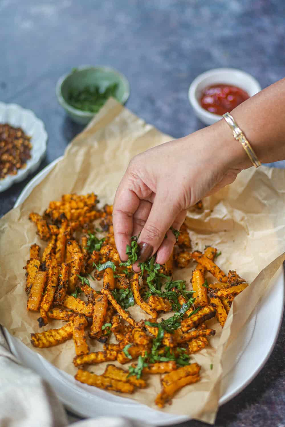 Hand sprinkling fresh cilantro over crispy air fryer butternut squash fries on parchment paper.