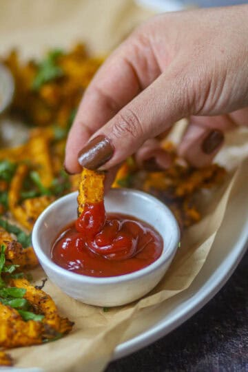 Close-up of dipping air fryer butternut squash fry into ketchup on a plate of crispy fries.