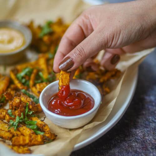 Close-up of dipping air fryer butternut squash fry into ketchup on a plate of crispy fries.