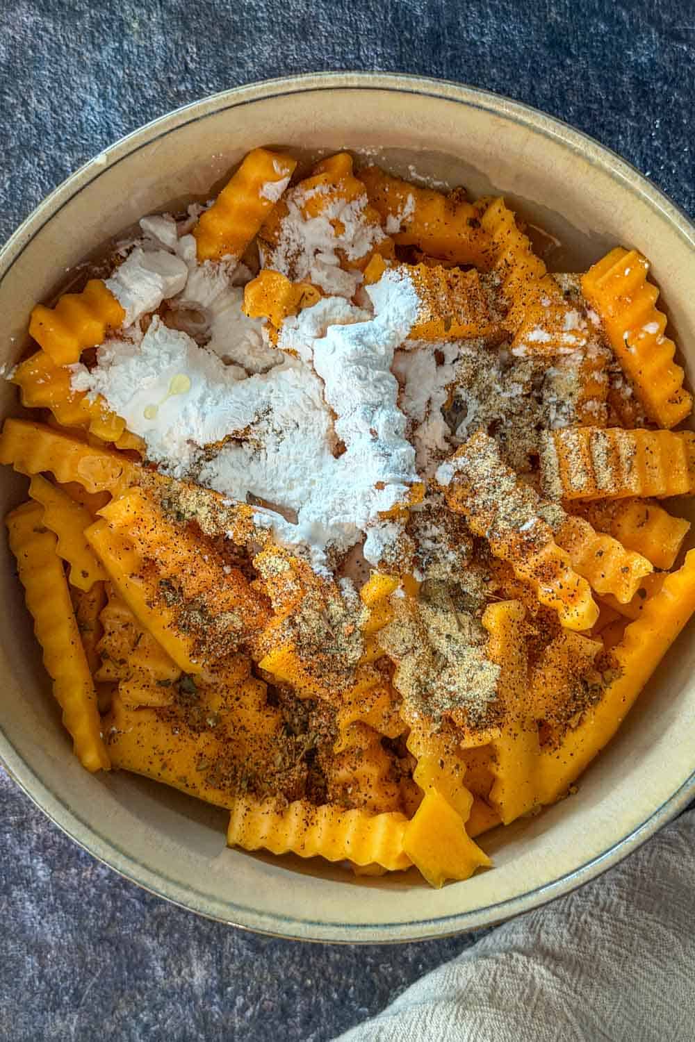 Raw crinkle-cut butternut squash fries in a bowl with spices and cornstarch added on top.