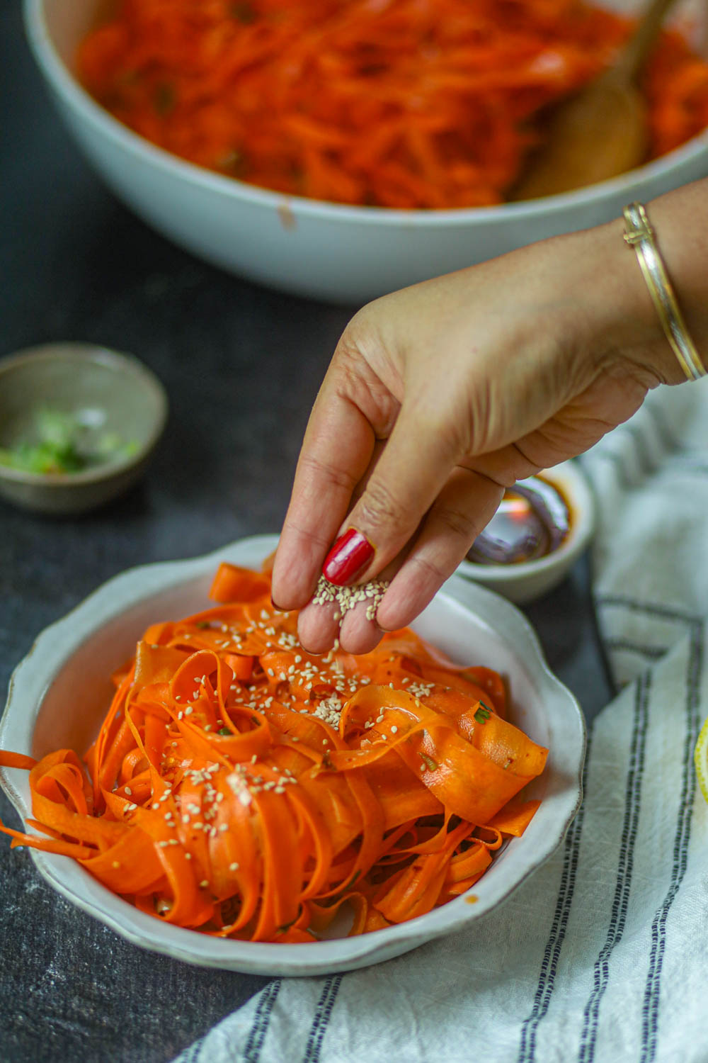 Hand sprinkling sesame seeds over carrot ribbon salad in a small bowl.