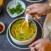 Hands holding a white bowl of creamy yellow lentil dal soup garnished with cilantro and roasted cumin.